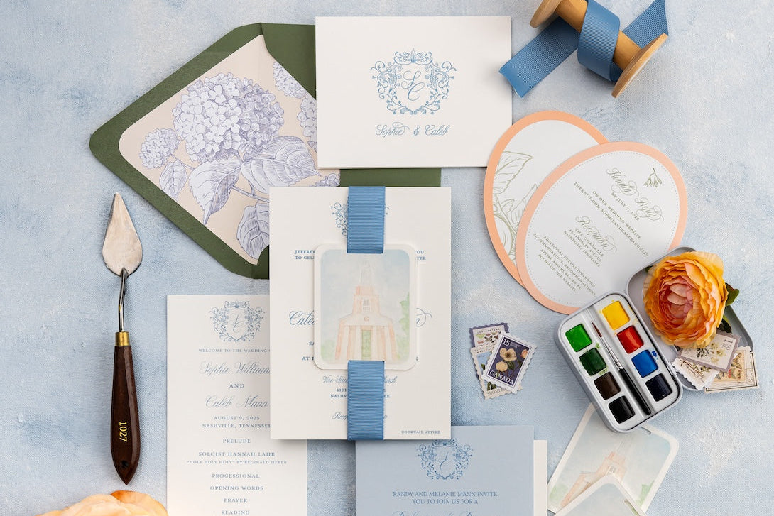 Letterpress custom wedding invitation suite with, watercolor venue tags, ribbons, round shaped cards, flowers and stationery items on a light blue surface